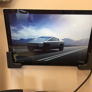 May include: A silver Tesla Cybertruck driving on a road with mountains in the background. The truck is on a computer screen mounted on a wall.