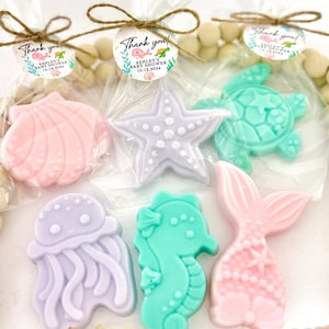 May include: A collection of sea-themed soaps in various shapes, including a seashell, starfish, turtle, jellyfish, seahorse, and mermaid tail. Each soap is a different pastel color and packaged in clear cellophane with a tag that says "Thank you!" and "Ashley's Baby Shower 10.12.2024".