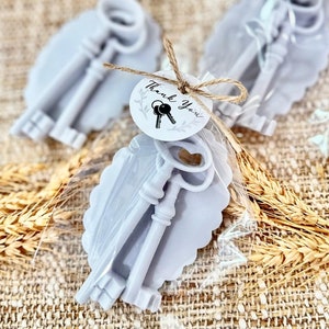 May include: Two silver-colored key-shaped soaps wrapped in clear cellophane with a small tag that says "Thank You" and a small image of keys. The soaps are on a light brown burlap background.