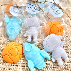 May include: A set of five astronaut and rocket themed party favors. The favors are made of soap and come in blue, white, and orange. The favors are packaged in clear cellophane bags with a tag that reads "Hope you had a blast - Theodore".