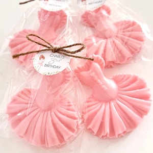 May include: Pink ballerina-shaped chocolate favors with a white circle tag that says "Charli's 3rd Birthday" in gold lettering. The favors are wrapped in clear cellophane bags and tied with twine.