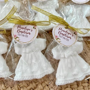 May include: White favour boxes with a white dress-shaped soap inside. Each box has a gold ribbon and a round tag with the text "Penelope's Baptism 12/18/22" in burgundy text with gold accents.