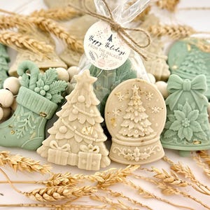 May include: A collection of sage green and cream-colored holiday-themed wax melts shaped like stockings, Christmas trees, snow globes, and bells. A clear plastic bag contains additional wax pieces with a tag reading Happy Holidays FROM THE WALKERS 2026.