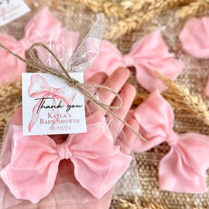 May include: Pink bow-shaped soap favors in clear cellophane bags tied with twine. A small card with the text "Thank you Kayla's Baby Shower 08/12/24" is attached to the bag.
