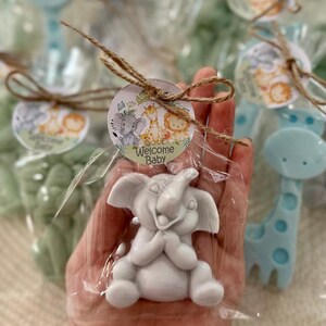 Puede incluir: Una peque&ntilde;a figurita de elefante blanco con una cara sonriente, empaquetada en una bolsa de pl&aacute;stico transparente con un lazo de cuerda marr&oacute;n y una etiqueta redonda que dice "Bienvenido Beb&eacute;". La etiqueta presenta un elefante, una jirafa y un le&oacute;n de dibujos animados en un entorno de selva.