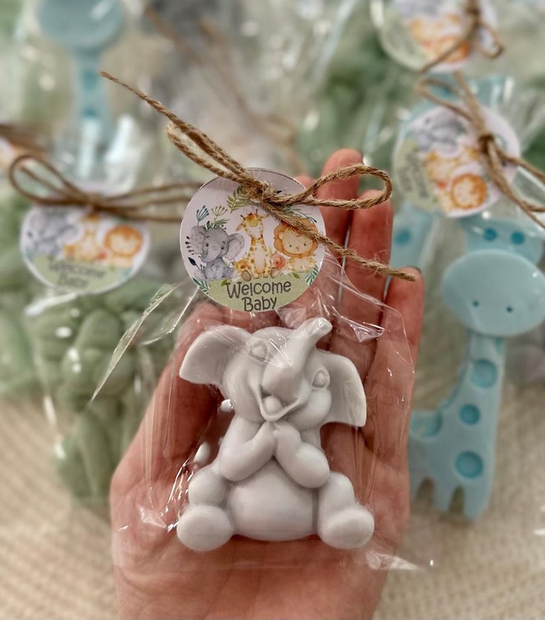 Puede incluir: Una peque&ntilde;a figurita de elefante blanco con una cara sonriente, empaquetada en una bolsa de pl&aacute;stico transparente con un lazo de cuerda marr&oacute;n y una etiqueta redonda que dice "Bienvenido Beb&eacute;". La etiqueta presenta un elefante, una jirafa y un le&oacute;n de dibujos animados en un entorno de selva.