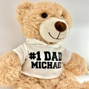 May include: A beige teddy bear wearing a white hoodie with the text "#1 DAD MICHAEL" printed in black.