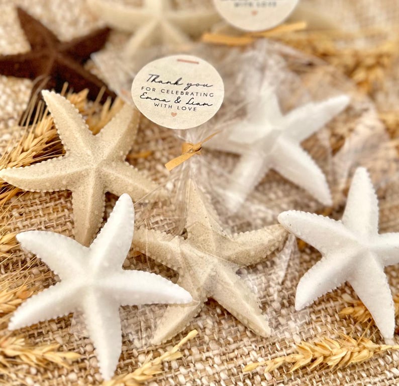 May include: White and beige starfish favors in clear cellophane bags tied with a brown ribbon. A round tag with the text "Thank you for celebrating Emma & Liam with love" is attached to the ribbon.
