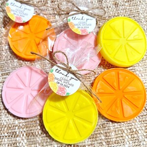 May include: A set of six favors for a bridal shower, each wrapped in cellophane and tied with twine. The favors are shaped like orange slices and are in three colors: pink, yellow, and orange. Each favor has a small tag attached with the text "Thank you Olmas Bridal Shower 08-16-2024".
