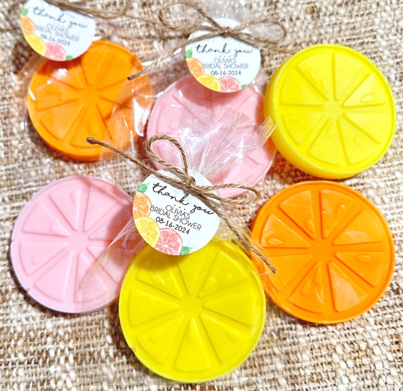 May include: A set of six favors for a bridal shower, each wrapped in cellophane and tied with twine. The favors are shaped like orange slices and are in three colors: pink, yellow, and orange. Each favor has a small tag attached with the text "Thank you Olmas Bridal Shower 08-16-2024".