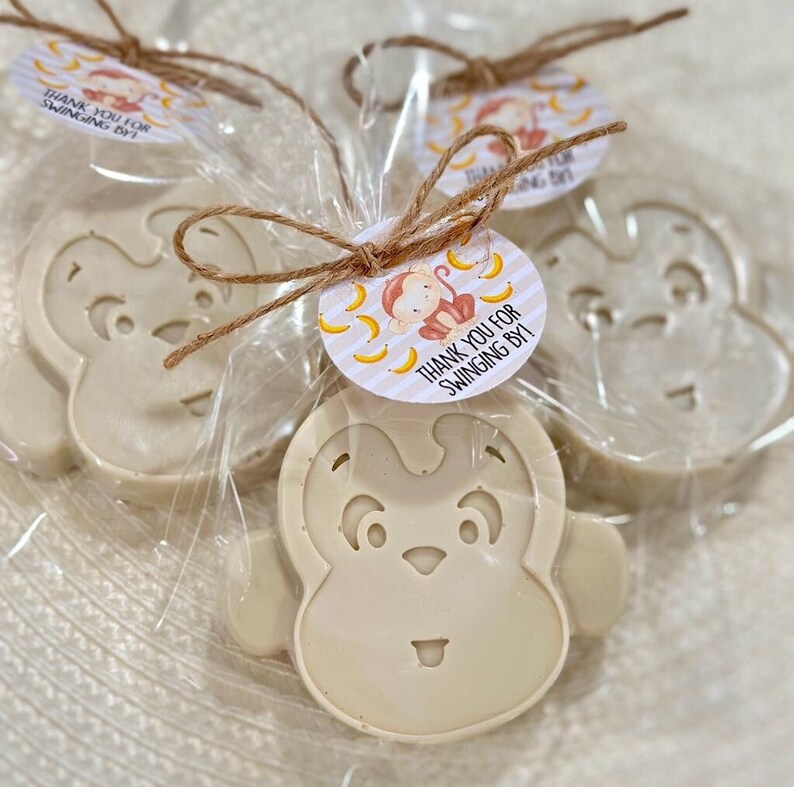 May include: Three small monkey-shaped soaps wrapped in clear cellophane bags with a brown twine bow. Each soap has a white monkey face with black eyes and a small mouth. Each bag has a round tag with a monkey and bananas on a striped background. The tag says "Thank you for swinging by!"