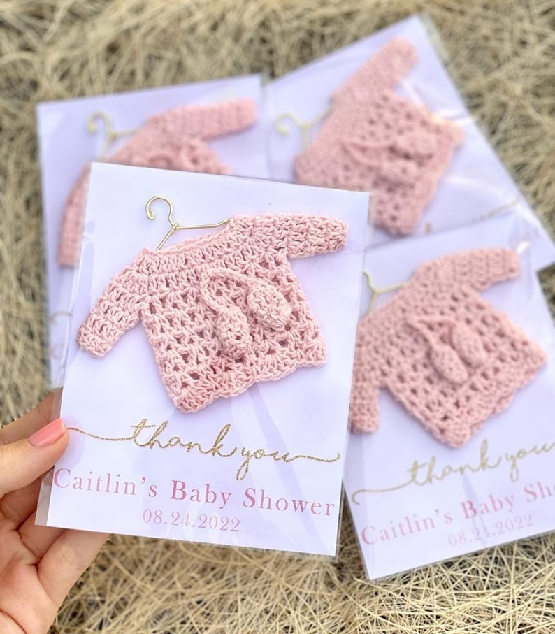 May include: Pink crocheted baby sweater with a hanger, packaged in a clear plastic bag with a white card that says "thank you" and "Caitlin's Baby Shower 08.21.2022".