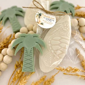 May include: A collection of tropical-themed party favors. The image features light green palm tree-shaped soaps, a surfboard-shaped soap with a turtle design, and a tag that reads "thank you for brightening our day Baby Taylor on Board."