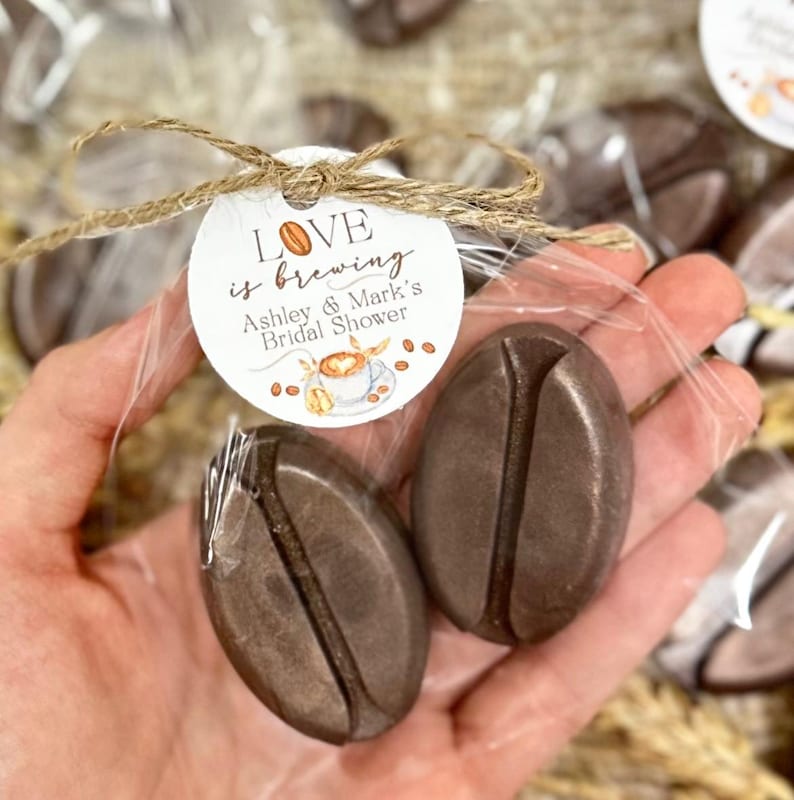 May include: Two brown coffee bean shaped soaps wrapped in clear cellophane with a brown twine bow and a round white tag that reads "Love is brewing Ashley & Mark's Bridal Shower".