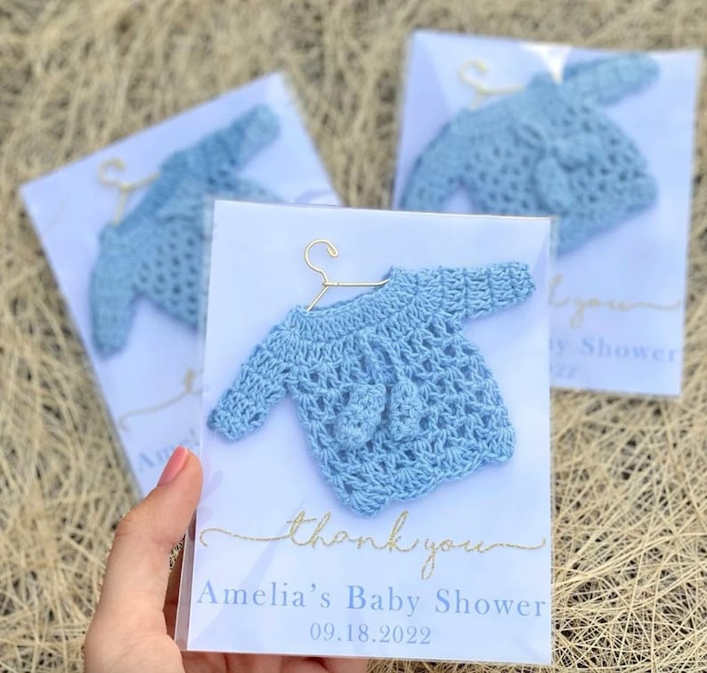 May include: Blue crocheted baby sweater on a hanger with a thank you card that says "Amelia's Baby Shower 09.18.2022"