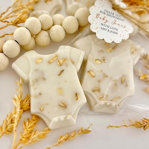 Op de afbeelding: Twee witte babyrompertjes in de vorm van traktaties met gouden accenten, verpakt in doorzichtig plastic. De traktaties zijn versierd met goudvlokken en liggen naast een rij houten kralen en tarwearen. Een label luidt "Bedankt voor het verwennen van Baby Jones met liefde - Amy & John."