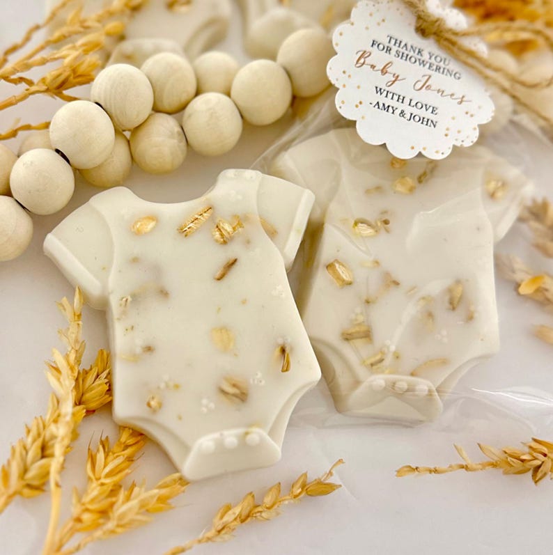 Op de afbeelding: Twee witte babyrompertjes in de vorm van traktaties met gouden accenten, verpakt in doorzichtig plastic. De traktaties zijn versierd met goudvlokken en liggen naast een rij houten kralen en tarwearen. Een label luidt "Bedankt voor het verwennen van Baby Jones met liefde - Amy & John."