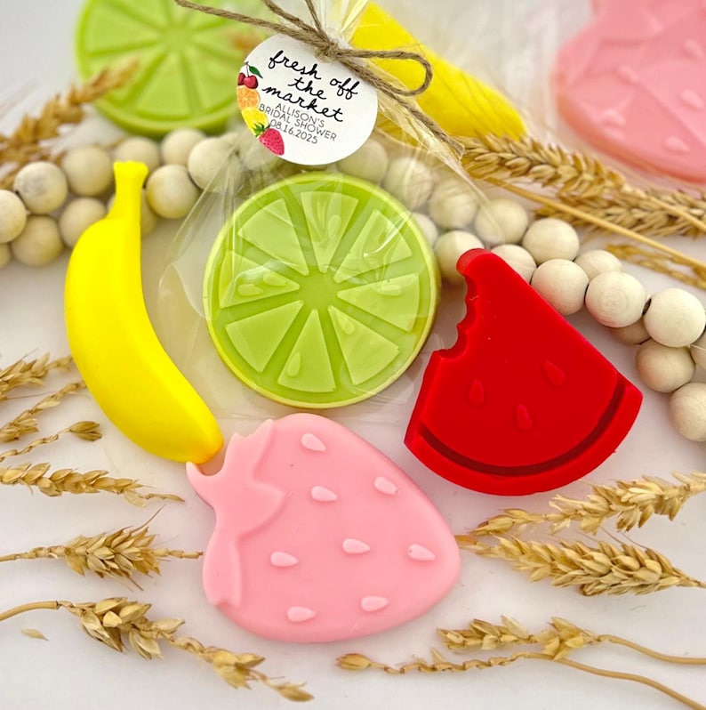 May include: A set of four colorful fruit-shaped soaps, including a lime, a banana, a strawberry, and a watermelon slice. The soaps are packaged in a clear plastic bag with a tag that reads "fresh off the market, Allison's Bridal Shower, Date 16.2023".