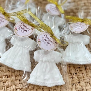 May include: White favour bags containing a white dress-shaped soap. Each bag is tied with a gold ribbon and features a round tag with the text "Penelope's Baptism 12/18/22" in pink and gold lettering.