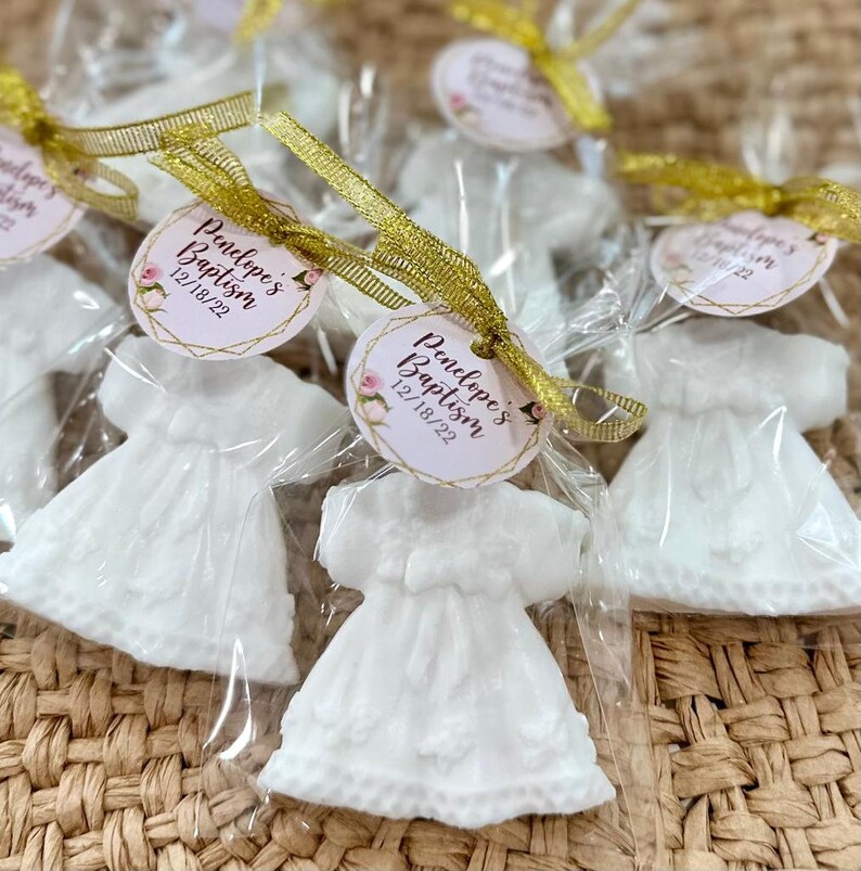 May include: White favour bags containing a white dress-shaped soap. Each bag is tied with a gold ribbon and features a round tag with the text "Penelope's Baptism 12/18/22" in pink and gold lettering.
