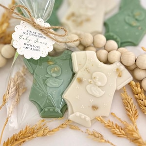 May include: Baby shower favors featuring onesie-shaped cookies in green and white, decorated with a duck design. The cookies are packaged in clear cellophane bags and tied with twine. A tag reads "Thank you for showering Baby Jones."