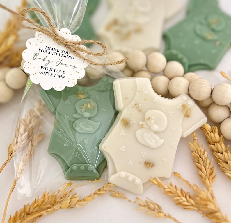 May include: Baby shower favors featuring onesie-shaped cookies in green and white, decorated with a duck design. The cookies are packaged in clear cellophane bags and tied with twine. A tag reads "Thank you for showering Baby Jones."