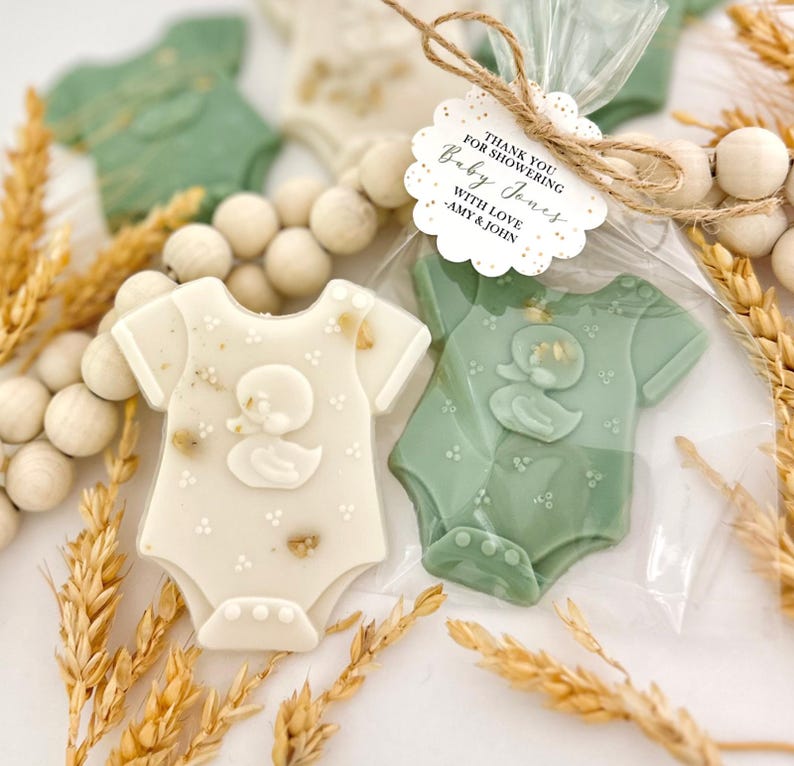 May include: Baby shower favors shaped like onesies, in white and sage green, with a duck design. The favors are packaged in clear cellophane bags and tied with twine. The tag reads "Thank you for showering Baby Jones with love, Amy & John."