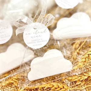 May include: White cloud-shaped favors with a round tag that reads "on cloud nine Ashley's Bridal Shower 09.12.2024".