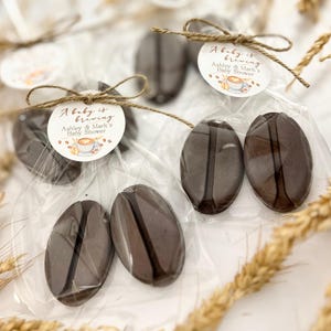 May include: Coffee bean-shaped favors in clear cellophane bags, tied with twine and a circular tag. The tag reads "A baby is brewing" and "Ashley & Mark's Baby Shower" with a coffee cup illustration. The favors are a dark brown color.