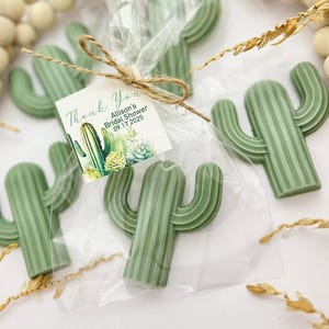May include: Green cactus-shaped favors, individually wrapped in clear cellophane and tied with twine. Each favor has a tag that reads "Thank You" and mentions a bridal shower date. The favors are arranged on a light-colored surface.