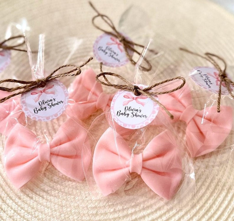 May include: Pink satin bows tied with twine and packaged in clear cellophane bags. Each bag has a round tag with the text "Olivia's Baby Shower" in white lettering on a pink and white polka dot background.