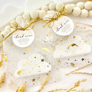 May include: Two cloud-shaped favors in sheer bags, adorned with gold stars and crescent moons. Each favor has a tag that reads "WE ARE ON cloud nine THANK YOU FOR SHOWERING BABY WALKER WITH LOVE 09.16.2025". The favors are tied with gold ribbon.