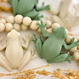 May include: Close-up of several frog figurines in shades of green and cream. The frogs are positioned near a string of wooden beads and dried wheat stalks. The frogs have outstretched limbs and are in a playful pose.
