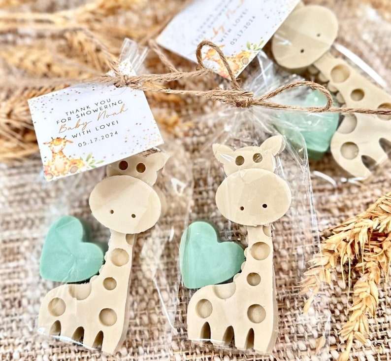 May include: Thank you for showering Baby Noah with love! 05.17.2024.  A set of two favor bags with a small giraffe-shaped soap and a heart-shaped soap in a light green color. The bags are tied with twine.