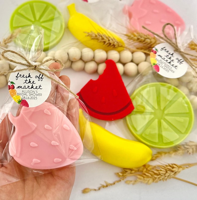 May include: A set of four fruit-shaped soaps wrapped in clear cellophane bags with a small tag that reads "fresh off the market Allison's Bridal Shower 08.16.2025". The soaps are shaped like a strawberry, a banana, a watermelon slice, and a lime.