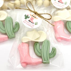 Op de afbeelding: Een doorzichtige plastic zak gevuld met kleine roze en groene cadeautjes. De cadeautjes zijn gevormd als cowboylaarzen, cactussen en cowboyhoeden. Een wit label met de tekst "Thank y'all for coming!" is aan de zak bevestigd.