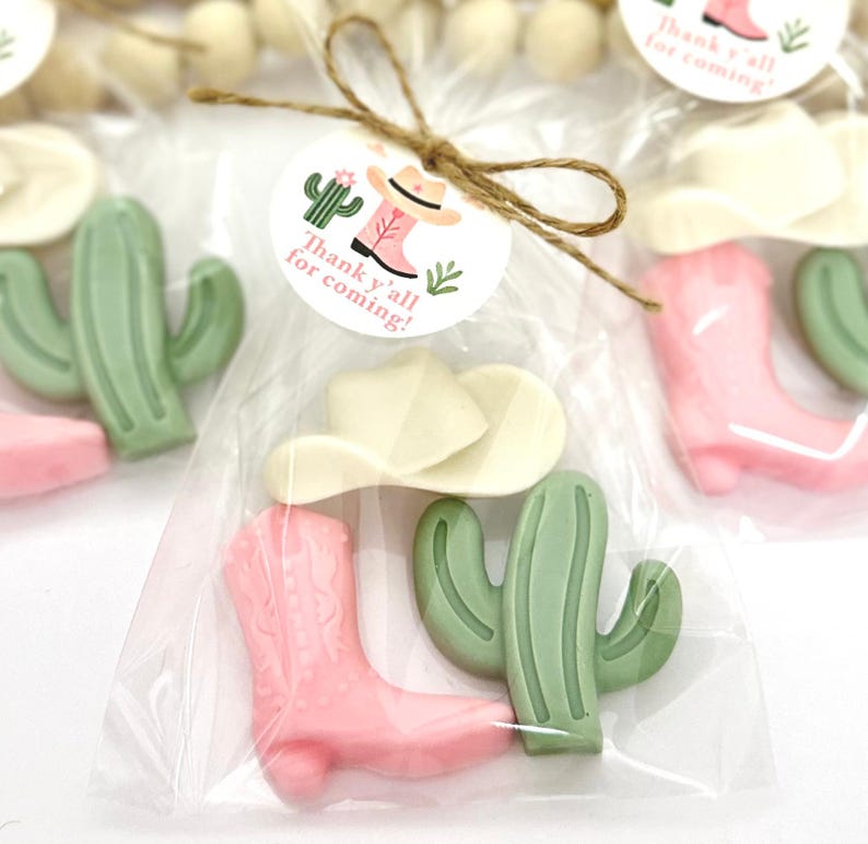 Op de afbeelding: Een doorzichtige plastic zak gevuld met kleine roze en groene cadeautjes. De cadeautjes zijn gevormd als cowboylaarzen, cactussen en cowboyhoeden. Een wit label met de tekst "Thank y'all for coming!" is aan de zak bevestigd.