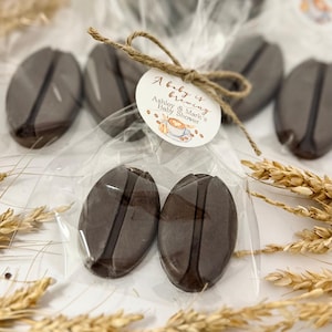 May include: Coffee bean-shaped favors in clear cellophane, tied with twine and a circular tag. The tag reads "A baby is brewing" with the names "Ashley & Mark's" and "Baby Shower" below a coffee cup illustration. The favors are dark brown.