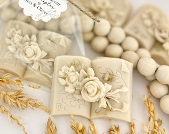 May include: Ivory-coloured, book-shaped soaps with floral designs. The soaps are embossed with roses and leaves. A tag reads "thank you for celebrating our new chapter Jess & Oliver". Wheat stalks and wooden beads are also in the image.