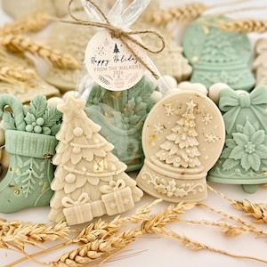 May include: A collection of holiday-themed wax melts in sage green and cream colors, featuring shapes of a Christmas tree, a stocking, a bell, and a snow globe. A clear plastic bag contains a wax melt with a tag reading Happy Holidays FROM THE WALKERS 2026.