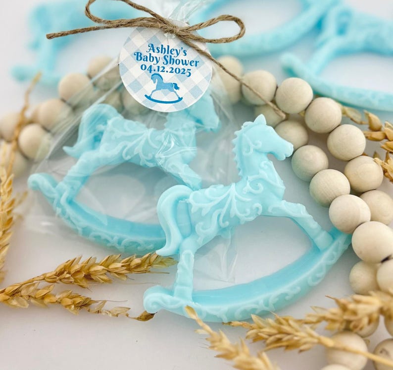 Może przedstawiać: Dwa niebieskie upominki w kształcie konika na biegunach z białą etykietą z napisem "Baby Shower Ashley 04.12.2025" i wzorem w niebiesko-białą kratkę.