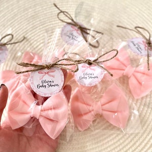 May include: Pink satin bows wrapped in clear cellophane bags with a brown twine bow and a round tag that says "Olivia's Baby Shower" in pink and white lettering.