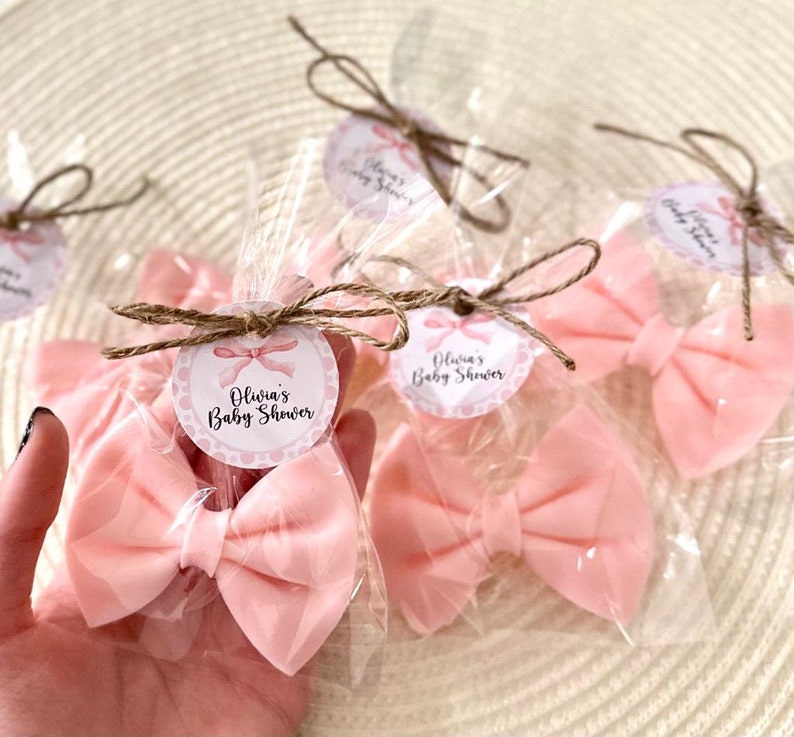 May include: Pink satin bows wrapped in clear cellophane bags with a brown twine bow and a round tag that says "Olivia's Baby Shower" in pink and white lettering.