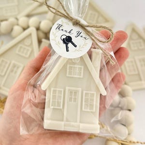 May include: A white house-shaped favor in a clear cellophane bag, tied with twine and a circular tag that reads "Thank You" with key graphics. The house has windows and a door.