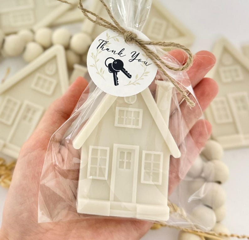 May include: A white house-shaped favor in a clear cellophane bag, tied with twine and a circular tag that reads "Thank You" with key graphics. The house has windows and a door.