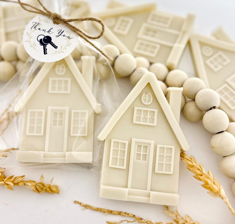 May include: Off-white house-shaped favors with detailed windows and doors, packaged in clear bags. A round tag with the words "Thank You" and a key illustration is attached with twine. Wooden beads and wheat stalks are also visible.