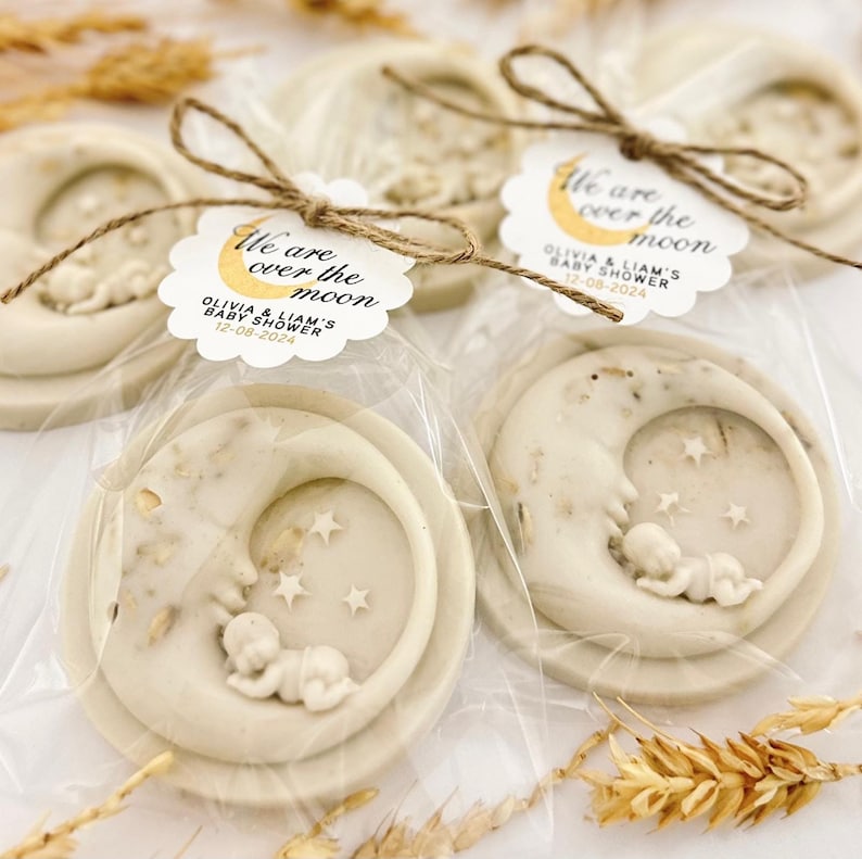 May include: Two white favor soaps shaped like a crescent moon with a baby sleeping inside. The soaps are wrapped in clear cellophane bags and tied with twine. Each soap has a round tag attached with the text "We are over the moon" and the date "12-08-2024".