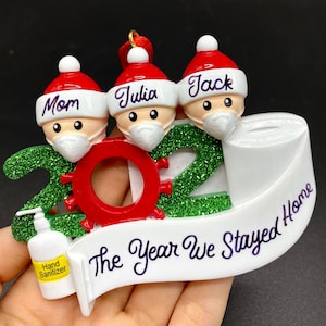 May include: A green and red 2021 Christmas ornament with three cartoon people wearing Santa hats and face masks. The ornament also features a roll of toilet paper, a bottle of hand sanitizer, and the text "The Year We Stayed Home".