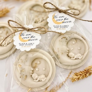 May include: Two white soap favors with a crescent moon design and a sleeping baby inside. Each soap is wrapped in clear cellophane and tied with twine. A white tag with gold lettering reads "We are over the moon Olivia & Liam's Baby Shower 12-08-2024".
