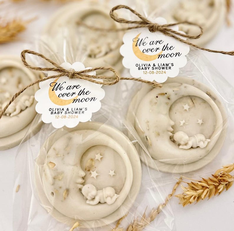 May include: Two white soap favors with a crescent moon design and a sleeping baby inside. Each soap is wrapped in clear cellophane and tied with twine. A white tag with gold lettering reads "We are over the moon Olivia & Liam's Baby Shower 12-08-2024".